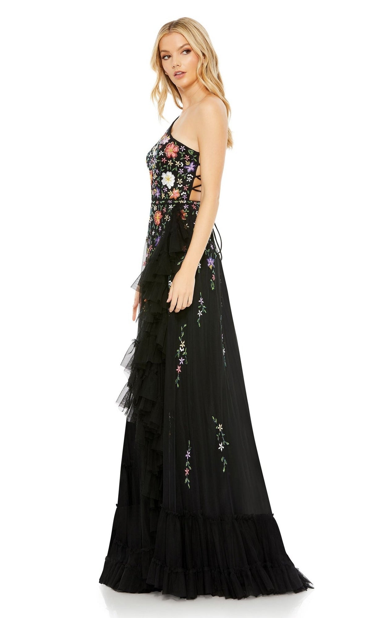 A woman poses in side profile wearing the Mac Duggal 9164 One-Shoulder Floral Formal Dress, featuring floral embroidery and tulle ruffles, against a plain white background.